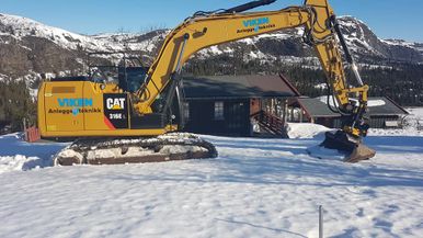 Gravemaskin i snø ved hytte
