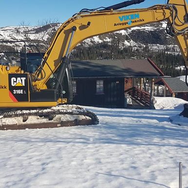 Gravemaskin i snø ved hytte
